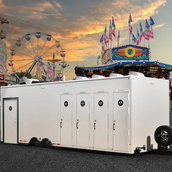 White multi-stall restroom trailer parked at outdoor carnival with ferris wheel and game booth in background at sunset