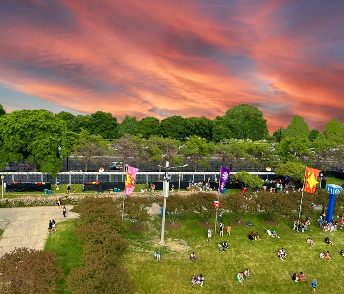Outdoor event with groups of people sitting on grass near colorful flags and tall trees under a vibrant sunset sky