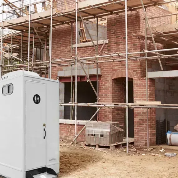 White portable restroom with ventilation window outside brick building under construction with scaffolding