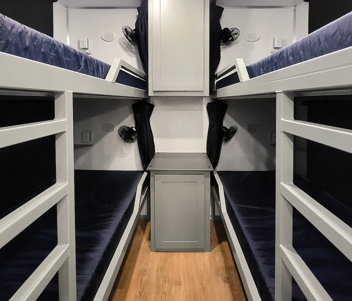 Four gray bunk beds with blue mattresses and individual fans in compact room with wood floor
