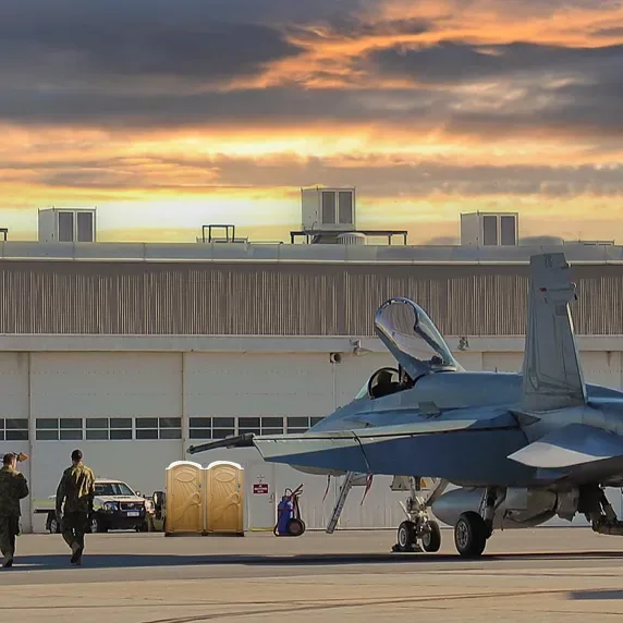 Two tan portable restrooms positioned near hangar with military jet and personnel at sunset