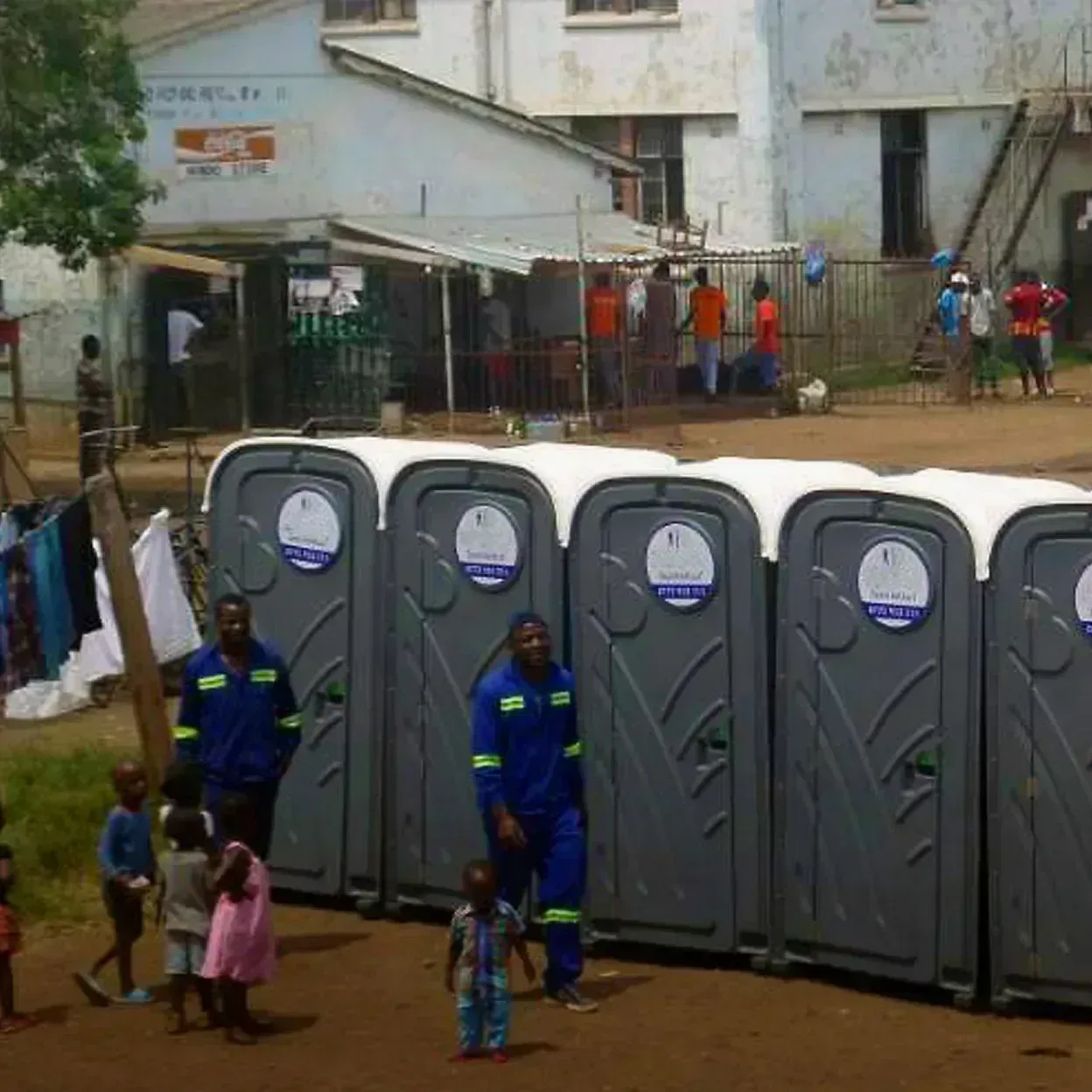 Row of gray portable restrooms with white roofs outdoors near building and people walking around