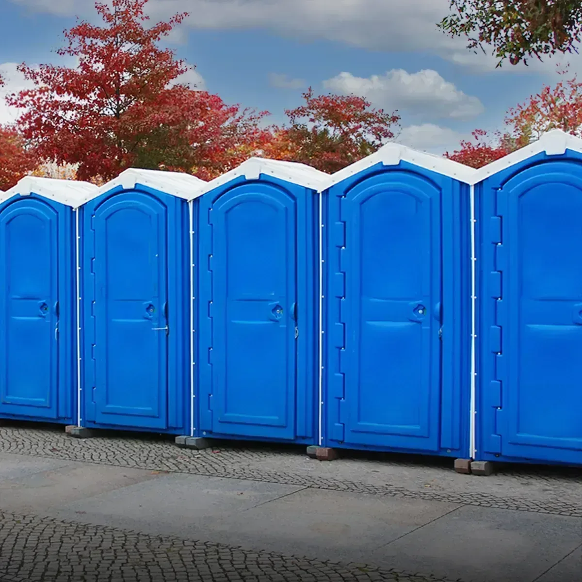 Row of blue portable restrooms with white roofs outdoors near autumn trees