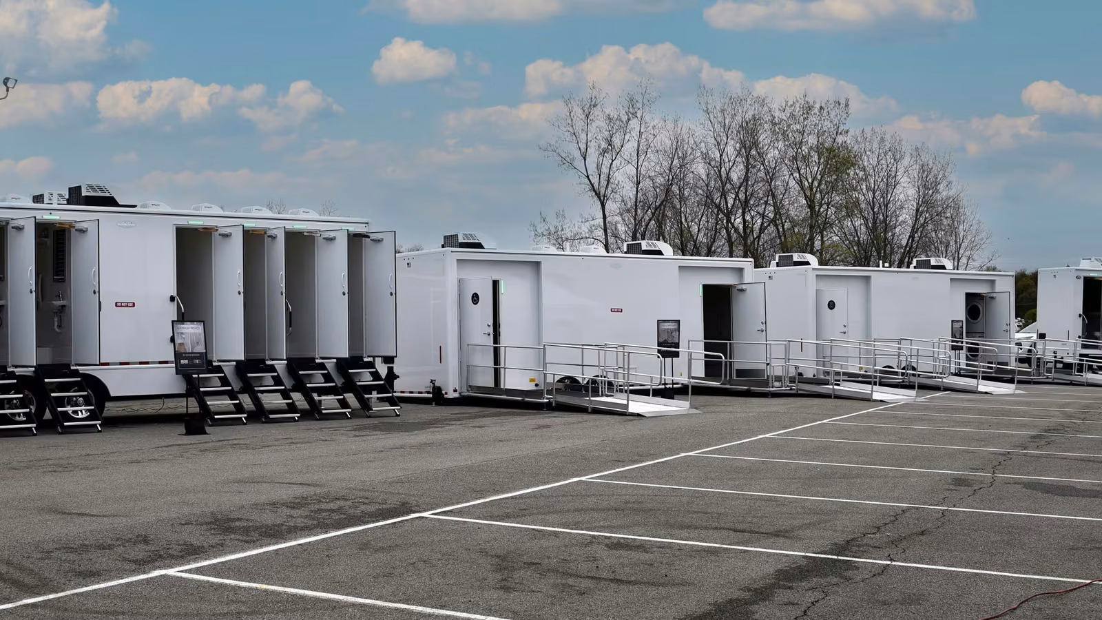 Row of portable restrooms and hygiene trailers in a parking lot under a cloudy sky.