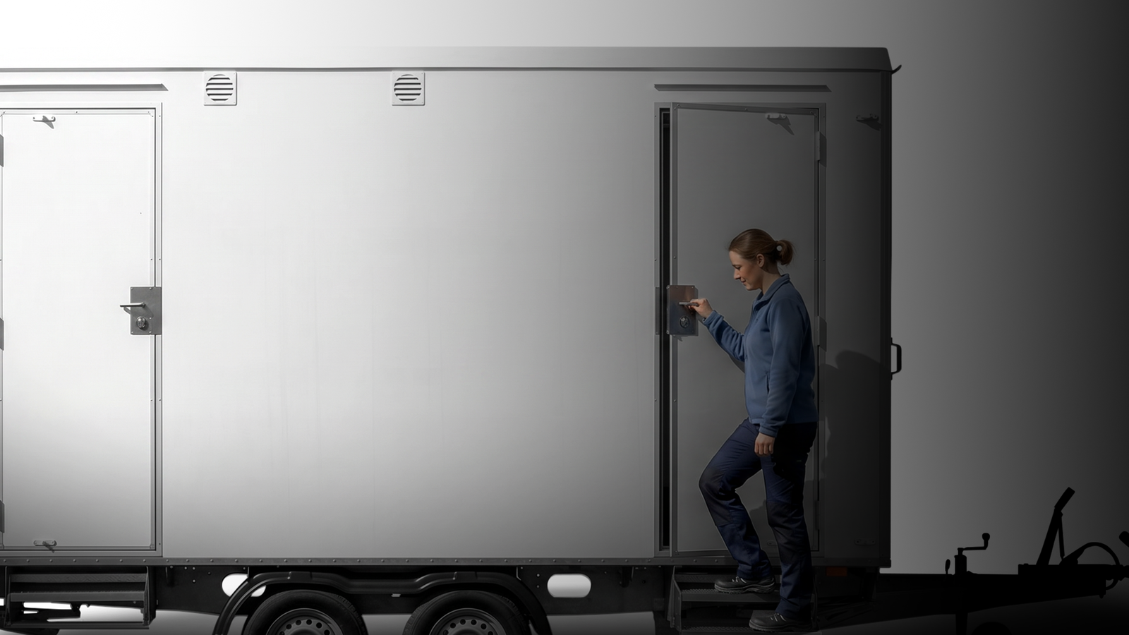 A person in a blue jacket is approaching a large, gray trailer, preparing to open a door.